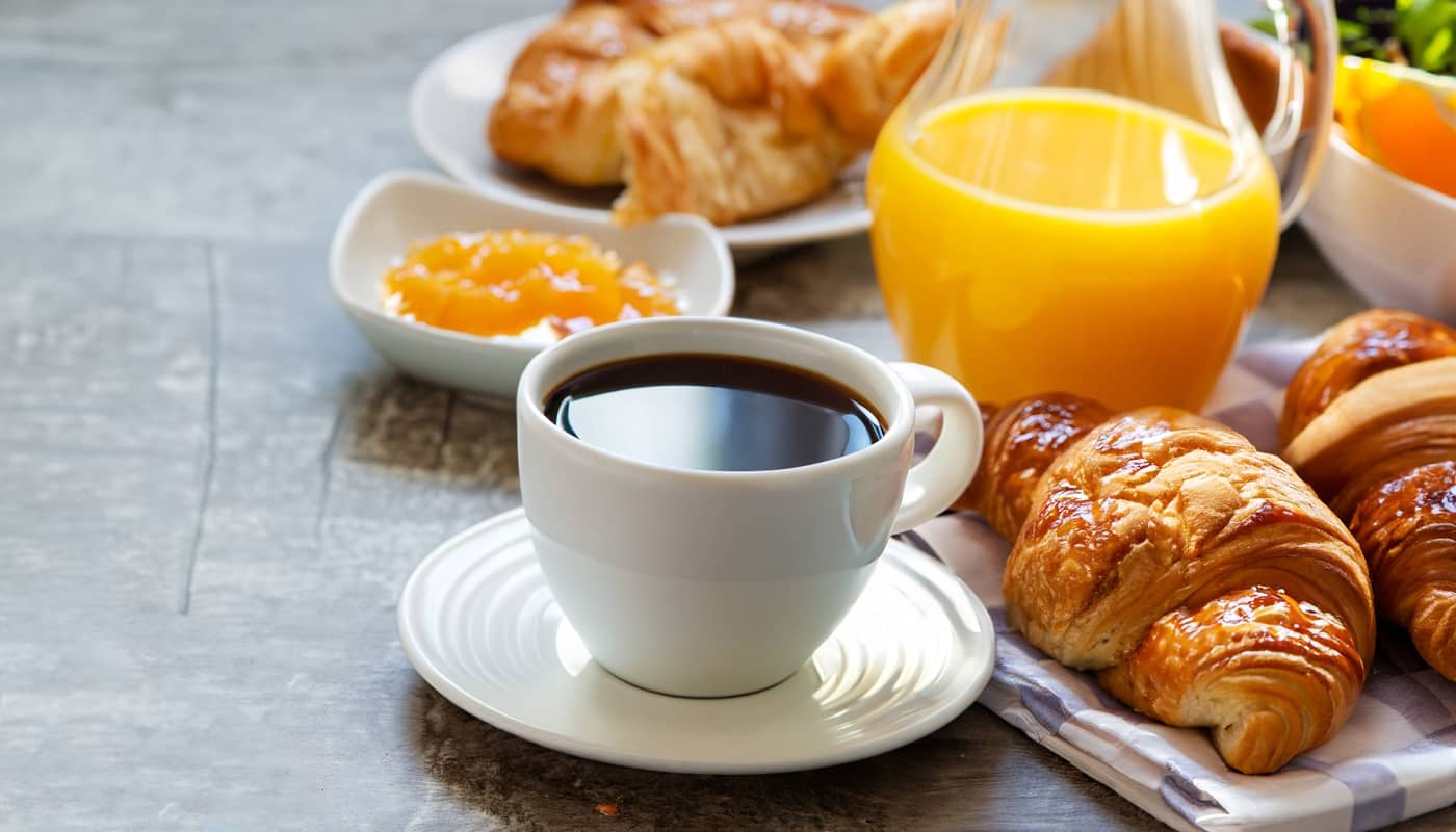 Das Bild zeigt eine Tasse Kaffee. Im Hintegrund sieht man frische Croissants, Orangensaft und Marmelade.